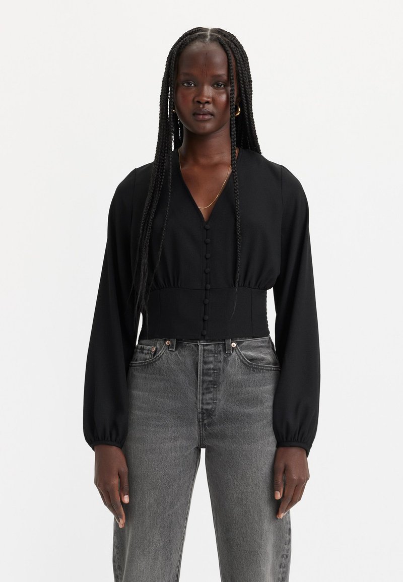 Schwarze Bluse mit langen Ärmeln und V-Ausschnitt, vorne mit Knopfdetails, kombiniert mit hochtaillierten grauen Jeans. Glatte Textur, taillierte Passform.