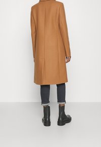 Lichtbruine wolblend jas, lang met een rechte snit, gedragen over donkere spijkerbroeken en zwarte enkellaarsjes. Opgerolde spijkerbroeken en minimale details.