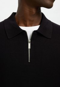 Gros plan d'une personne portant une chemise noire zippée avec une fermeture éclair argentée partiellement ouverte sur un sous-vêtement blanc.