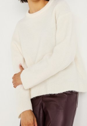 Osoba nosící měkký bílý pletený svetr a tmavě hnědé kožené kalhoty, s jednou rukou jemně držící opačné rameno.