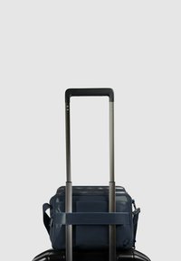 Bolso de viaje azul marino hecho de material brillante, con cierre de cremallera y una correa ajustable, colocado sobre una maleta oscura con ruedas.