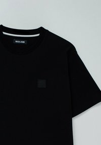 Camiseta negra de manga corta hecha de tela texturizada, con un pequeño parche de logo negro en el frente y un diseño de cuello redondo acanalado.