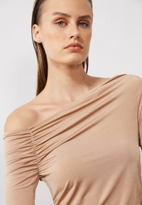 Beež off-shoulder pluus, millel on kurrulisus, valmistatud pehmest elastse kangast, millel on pikad varrukad ja pehme tekstuur.