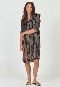 Zwarte jurk met verticale bruine strepen, korte mouwen en een kraag. Lichtgewicht stof met een vloeiende uitstraling, knielange snit, gedragen met slippers.