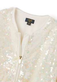 Giacca bianca per bambini con paillettes iridescenti, chiusura con zip e collo rotondo. L'etichetta mostra "OVS kids", taglia 8Y e paese di origine.