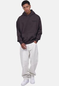 Brązowy hoodie z kangurzą kieszenią i wytłoczonym logo, w połączeniu z białymi, postarzanymi spodniami szerokimi i białymi sneakerami.