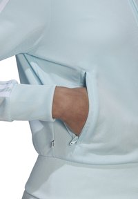 La mano derecha insertada en el bolsillo lateral con cremallera de una blusa de manga larga texturizada de color azul claro, con puños acanalados y sutiles rayas blancas en la manga.