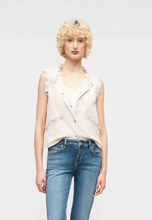 Jong persoon met kort krullend blond haar draagt een mouwloos lichtbeige vest met ruches en een blauwe spijkerbroek, kijkt recht vooruit tegen een effen achtergrond.