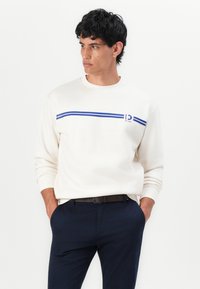 Crew-neck sweatshirt in cream with blue horizontal stripes across the chest and logo detail. Worn with navy trousers and a black belt.