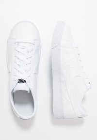 Vita lädersneakers med en texturerad vit sula, platt snördesign och en liten svart logotyp på tungan. Sidan har en slät swoosh.