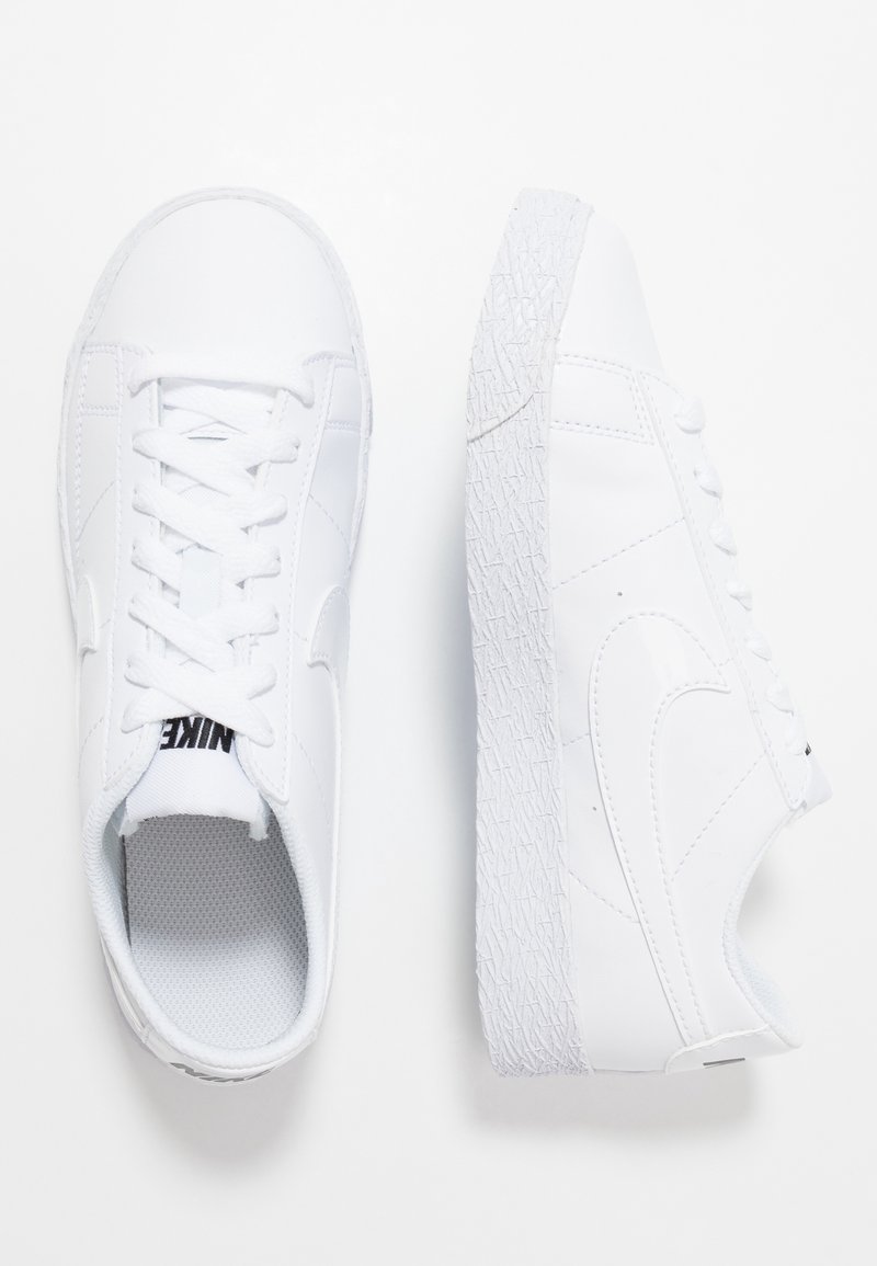 Vita lädersneakers med en texturerad vit sula, platt snördesign och en liten svart logotyp på tungan. Sidan har en slät swoosh.