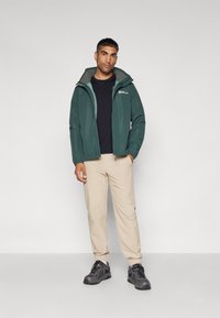 Groene waterdichte jas met capuchon, zwarte T-shirt, beige broek en grijze schoenen met zwarte accenten. Model staat tegen een effen achtergrond.