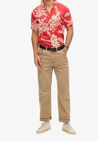 Camisa de manga corta roja con estampado floral y hojas blancas, combinada con pantalones color beige claro y zapatillas beige, con cinturón y reloj negros.