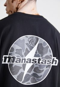 Schwarzes T-Shirt mit einem großen kreisförmigen Grafikdesign, das topografische Linien in Grau zeigt, sowie einem zentralen Blitzsymbol und dem Text "manastash" darunter.