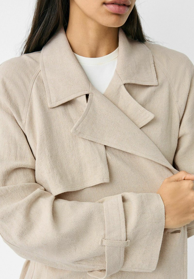 Cappotto beige strutturato con ampio colletto, design doppiopetto e polsini cinturati, realizzato in un morbido tessuto intrecciato.