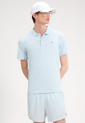 Giovane uomo con polo azzurro chiaro, pantaloncini abbinati, cappellino da baseball bianco e occhiali, in piedi con le mani dietro la schiena.