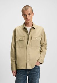 Beige button-up shirt made of sturdy fabric, featuring two front pockets with flaps and a classic collar, worn over a white T-shirt.