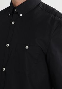 Camicia nera con bottoni realizzata in cotone, caratterizzata da un colletto a bavero, maniche lunghe e una tasca sul petto con bottoni bianchi.