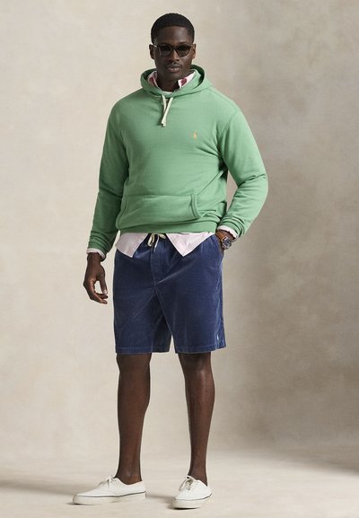 Sweatshirt verde com cordão, usado por cima de uma camisa rosa com botões. Calções de bombazina azul-marinho e ténis brancos completam o conjunto.