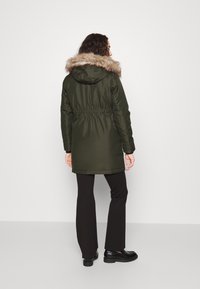 Parka verde oliva con capucha forrada de pelo, cintura ajustada y mangas largas. Combinada con pantalones negros de campana y zapatos negros gruesos.