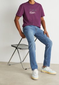 Lila bomull T-shirt med "Calvin Klein"-logotyp, blå denimjeans och vita sneakers med gröna och gula accenter, sittande på en metallstol.