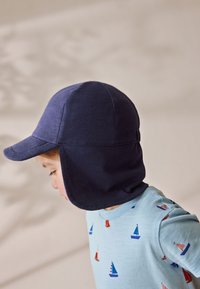 Gorra de marinero con orejeras hecha de tela suave, con una visera curva y detalles de costuras. La lleva puesta un niño con una camiseta azul claro con estampados de barcos.