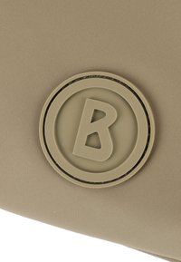 Accessorio in tessuto beige caratterizzato da un logo rotondo tattile con una "B" stilizzata al centro, superficie testurizzata e senza parti metalliche visibili.