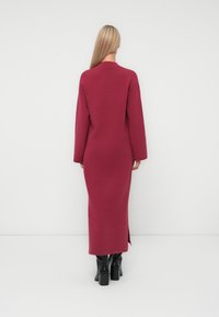 Lungo vestito bordeaux con maniche lunghe, una vestibilità comoda e spacchi laterali, abbinato a stivali neri al ginocchio su uno sfondo chiaro.