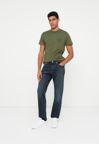 Olíva zöld t-shirt nyomtatott szöveggel, sötét farmernadrággal párosítva. A modellen fehér sportcipők vannak. Egyszerű dizájn, laza szabás, nincs figyelemfelkeltő minta.