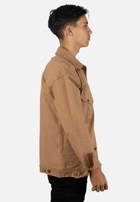 Chaqueta de mezclilla marrón claro con diseño clásico, bolsillo frontal en el pecho, cierre de botones y costuras visibles. Textura suave y ajuste ligeramente holgado.