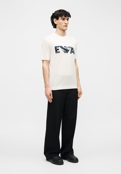 Νέος άνδρας όρθιος, φορώντας λευκό T-shirt με το κείμενο "EVA" και μαύρο παντελόνι φαρδιάς γραμμής, σε απλό ανοιχτό φόντο.