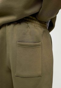 Olijfgroene sweatpants met een gladde textuur, elastische tailleband en een enkele steekzak aan de rechterkant. Eenvoudig, functioneel ontwerp.