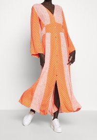 Longue robe orange avec des panneaux à motifs de pois et de fleurs, col en V et manches ballon. Portée avec des baskets blanches.