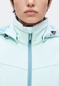 Gros plan d'une personne portant une veste bleu clair avec un col haut et une fermeture éclair bleue centrée.