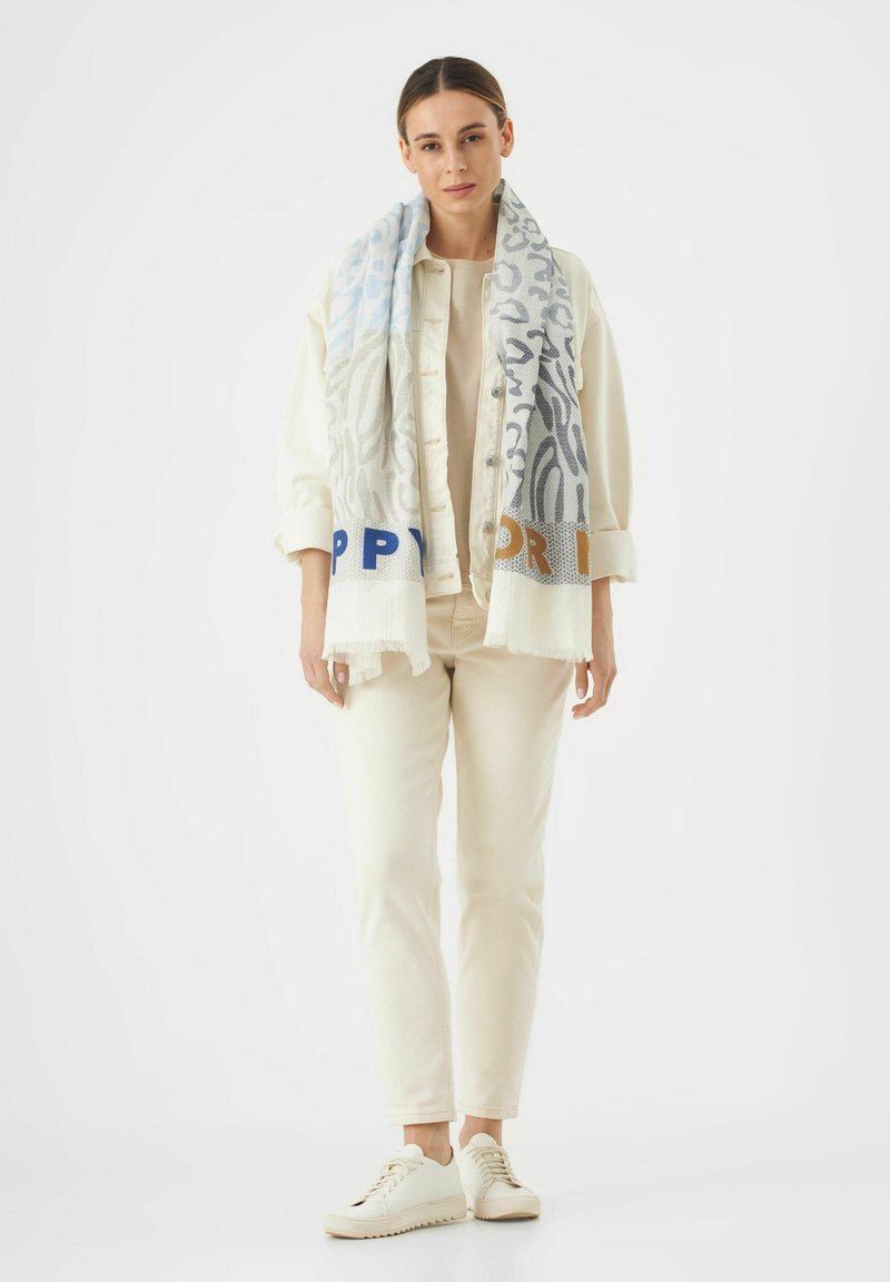 Foulard léger à motifs en bleu, gris et crème, avec le texte "HAPPY" en bleu et orange. Le mannequin porte une tenue neutre et des baskets.