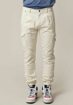 Crème cargobroek met een slim fit, voorzien van zijzakken, een ritsluiting en elastische boorden, gecombineerd met witte en marineblauwe sneakers.