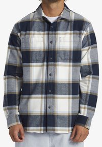 Rutigt flanellskjorta i marinblått, beige och vitt. Har en knäppbar krage, två bröstfickor och långa ärmar med knäppte manschetter.
