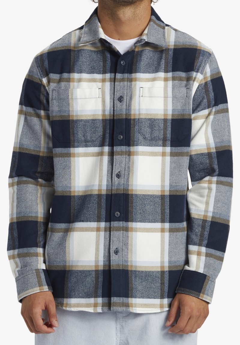 Rutigt flanellskjorta i marinblått, beige och vitt. Har en knäppbar krage, två bröstfickor och långa ärmar med knäppte manschetter.
