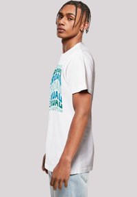 Weißes T-Shirt mit mehrfarbigem, fett gedrucktem "JORDAN"-Text in geschichteter Gestaltung. Kurze Ärmel und entspannte Passform. Getragen mit hellblauen Jeans.