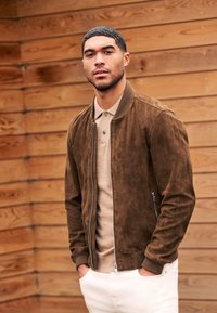 Junger Mann mit kurzen schwarzen Haaren, der eine braune Wildlederjacke, ein beiges Poloshirt und weiße Hosen trägt, steht vor einer Holzwand.
