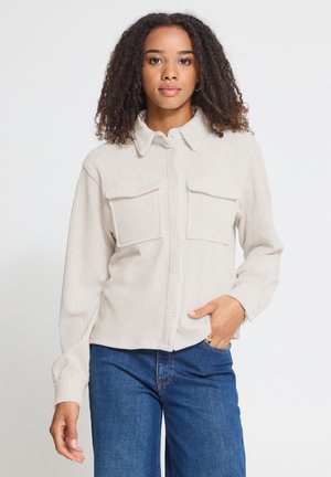 Femme aux cheveux bouclés portant une chemise beige clair boutonnée avec des poches sur le devant et un jean bleu, debout sur un fond blanc uni.