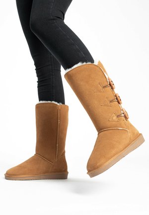 Tan suede knee-high boots with white faux fur lining, featuring three triangular accents and wooden buttons on the side, set against a white background.