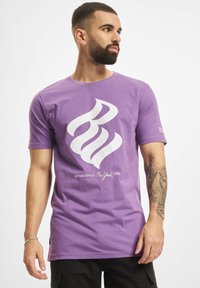 Fialové tričko s krátkým rukávem a bílým grafickým designem, obsahující stylizovaný text a logo, vyrobené z bavlny. Obsahuje kulatý výstřih.