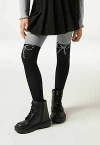 Sorte og grå knæhøje leggings med rhinsten-bue detaljer, parret med sorte snørrede kampstøvler og en plisseret grøn nederdel.