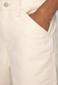 Pantalones cortos de algodón beige con una textura suave, que cuentan con bolsillos laterales y detalles de costura. Cierre de botón metálico en la cintura.