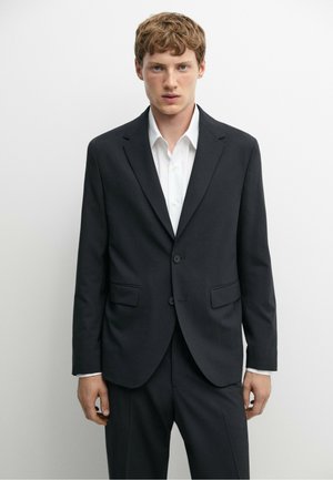 Jeune homme aux cheveux courts roux portant une veste de costume noire sur une chemise blanche, debout devant un fond clair uni.
