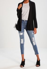 Blazer negro sobre una camiseta gris, combinado con jeans azules desgastados que presentan grandes desgarros y un dobladillo deshilachado. Mules negros con tacón completan el look.