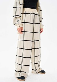 Ruime beige broek met zwart ruitpatroon, zachte stof, elastische tailleband en zijsplitten. Gecombineerd met donkere instappers.