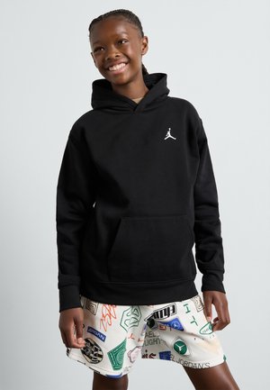 Schwarzer Hoodie mit Kängurutasche, ausgestattet mit einem weißen Jordan-Logo auf der linken Brust; das Modell trägt hellfarbene, gemusterte Shorts.