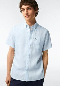 Camicetta azzurra chiara a maniche corte con bottoni, realizzata in lino, dotata di una tasca sul petto e un piccolo logo Lacoste ricamato in verde scuro.